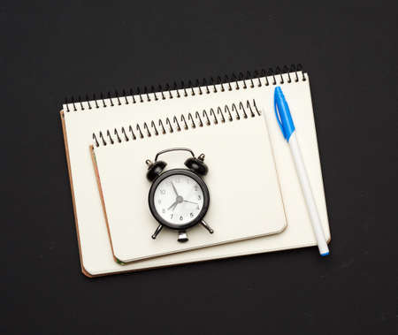 black round clock and stack of spiral open notebooks with blank white sheets on a black background, top viewの写真素材