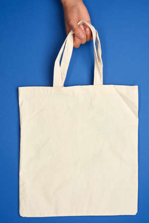 female hand holds an empty beige textile bag by the handles on a blue background, rejection of plastic bags, zero wasteの写真素材