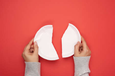 two female hands holding halves of a plastic disposable white plate on a red background, avoiding...
