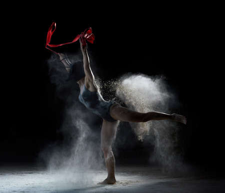 beautiful caucasian woman in a black bodysuit with a sports figure dancing with red ribbon on a black background, gymnastic exercises in flying white flourの写真素材