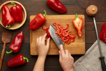 woman cuts red bell pepper into pieces on wooden board, brown wooden table, top view