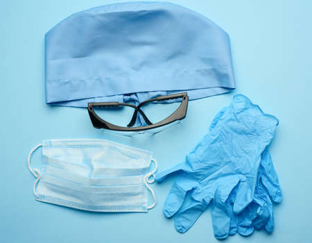 textile blue cap, disposable medical mask, pair of gloves and plastic glasses on a blue background, top view. Medic protective clothingの写真素材