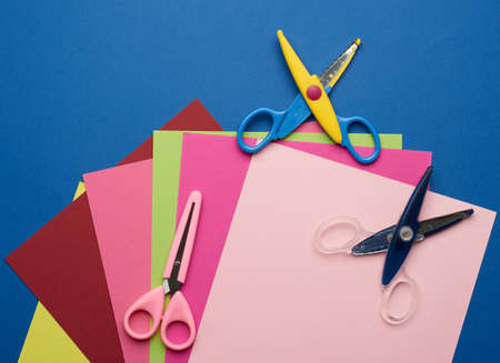 multicolored craft paper and plastic scissors on blue background, top view