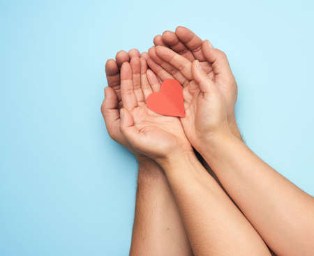 two female hands lie in male palms and hold a red paper heart on a blue background, top view....