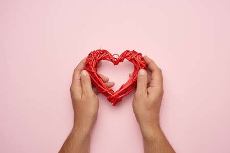 two male hands holding a red wicker heart on a pink background, love concept