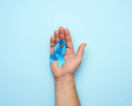 male hand holds blue silk ribbon in the shape of a loop on a blue background, symbol of the fight...