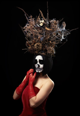 woman of Caucasian appearance with skeleton make-up stands in a red velor dress with a large crown of dry branches, black backgroundの写真素材