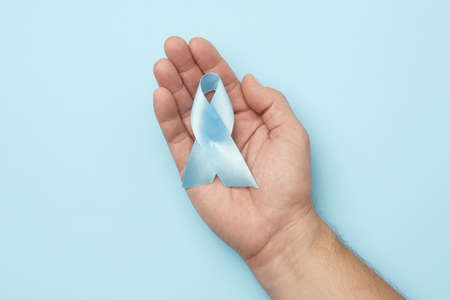 male hand holds blue silk ribbon in the shape of a loop on a blue background, symbol of the fight and treatment of prostate cancer, top viewの写真素材