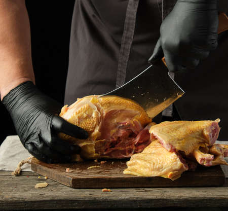 chef in black uniform and latex gloves chopping raw chicken into pieces on a brown wooden board, cooking processの写真素材