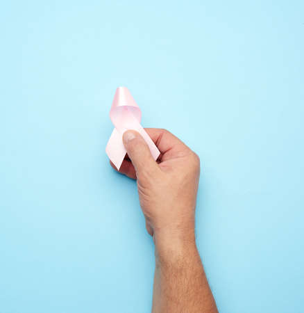 male hand holds a pink bow-shaped ribbon on a blue background. Breast cancer awareness campaign...