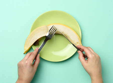 round green ceramic plate with a whole banana, two hands hold a knife and fork on a green...
