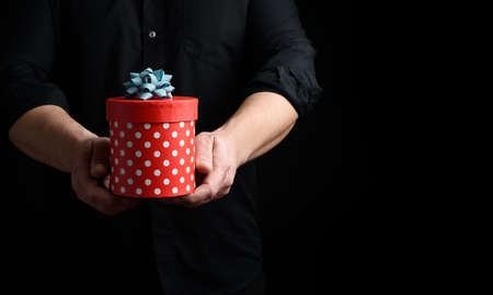adult man in a black shirt holds in his hand a round red box of polka dots with a blue bow on a...