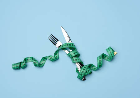 fork and knife wrapped in green measuring tape on blue background, weight loss concept, flat layの写真素材