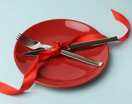 round plate and metal fork and knife tied with a red silk ribbon, blue background, place for an inscription, top viewの写真素材