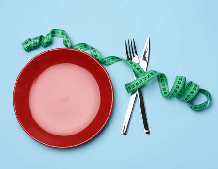 red round plate and fork and knife wrapped in green measuring tape on blue background, weight loss concept, flat layの写真素材