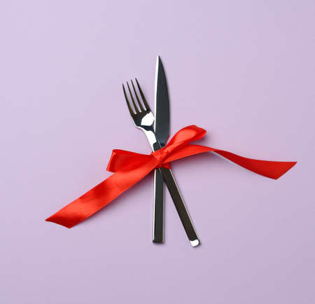 metal fork and knife tied with a red silk ribbon, purple background, top view