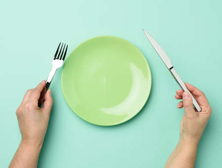 two hands hold a metal knife and fork on the surface of a round empty green plate, top viewの写真素材