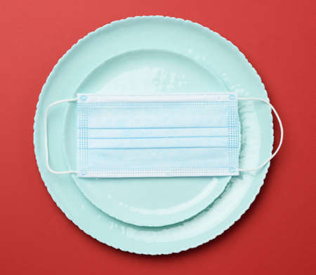 empty round green d ceramic plate on a red background and disposable medical mask on top, restaurant...