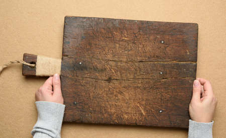 two female hands hold an empty old brown rectangular wooden cutting board, brown background, top viewの写真素材