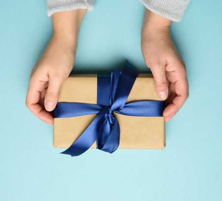 two female hands in a sweater hold a square gift box with a blue bow on a blue background, top view, festive backdropの写真素材