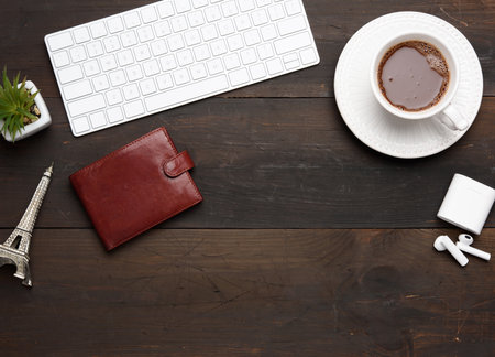 white wireless keyboard and wireless headphones and brown leather wallet on a wooden brown table, next to a white cup with coffee, top viewの写真素材
