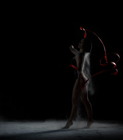 beautiful caucasian woman in a black bodysuit with a sports figure dancing with red ribbon on a black background, particles of flour fly in different directionsの写真素材