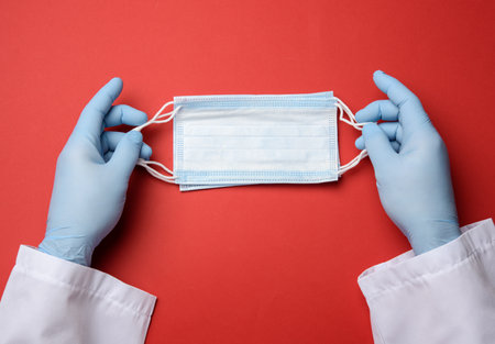 two male hands in blue latex gloves hold a medical disposable mask on a red background