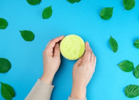 female hands of a young girl with smooth skin and round jar with cream, blue background with green leaves of raspberry, concept of spa treatments for the skinの写真素材