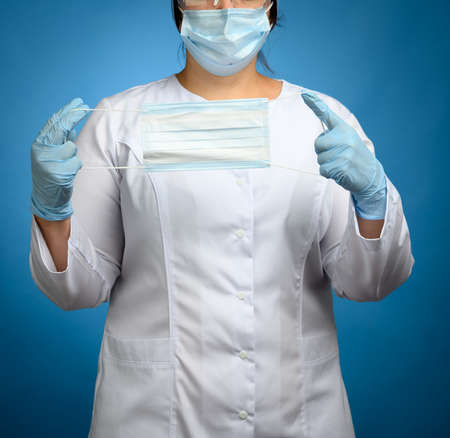 a woman doctor in a white medical coat, a disposable mask, protective plastic glasses and a cap stands on a blue background and holds a face mask, close upの写真素材