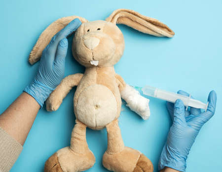 plush rabbit with a bandaged paw and two female hands of a doctor injecting an injection on a blue...