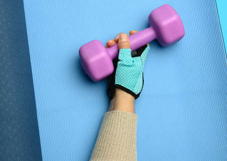 female hand in a blue sports glove holds a purple one kilogram dumbbell on a blue background, close upの写真素材