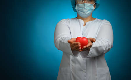 female medic in a white coat, a mask stands and holds a red heart on a blue background, the concept of donation and kindnessの写真素材