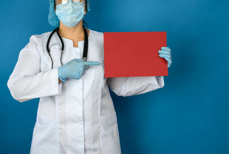 woman doctor in a white medical gown, a disposable mask, protective plastic glasses and a cap stands on a blue background and holds a blank red sheet of paperの写真素材