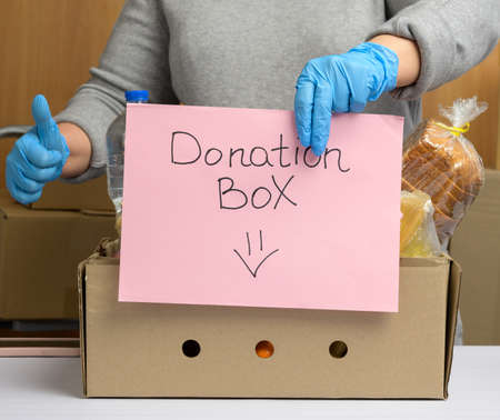 woman in gloves holds a sheet of paper with a lettering donation box and a cardboard box with food...