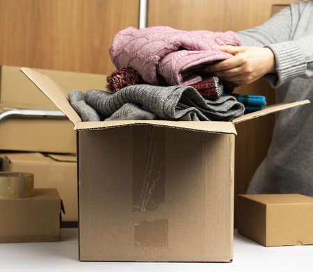 woman in a gray sweater collects clothes in a box, concept of assistance and volunteering, moving. Behind a stack of brown boxesの写真素材
