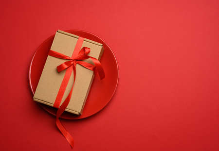 rectangular brown cardboard box tied with a silk red ribbon lies in a round ceramic red plate, top...