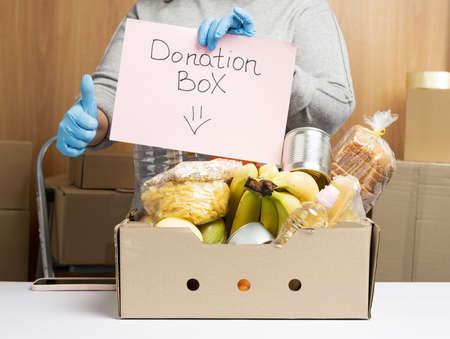 woman in gloves holds a sheet of paper with a lettering donation box and a cardboard box with food and things to help those in need, volunteering help conceptの写真素材