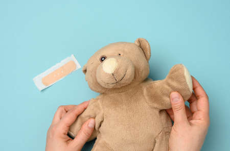 female hand holding a brown teddy bear with a torn off eye on a blue background, top viewの写真素材