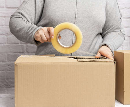 woman in a gray sweater holds a roll of duct tape and packs brown cardboard boxes. Moving conceptの写真素材