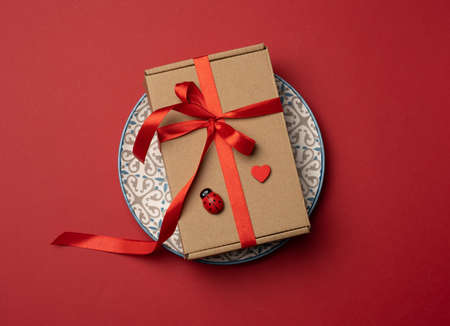 rectangular brown cardboard box tied with a silk red ribbon lies in a round ceramic red plate, top viewの写真素材