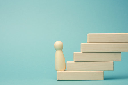 a wooden figurine of a man stands on a staircase made of blocks on the first step. The concept of achieving the set goals in business, career growth, start-up. Blue backgroundの写真素材