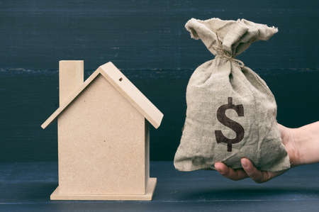 hand holds a canvas bag full of money and a wooden house on a blue background. The concept of buying real estate, renting out. Profitable investment in real estateの写真素材