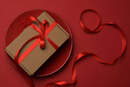rectangular brown cardboard box tied with a silk red ribbon lies in a round ceramic red plate, top...