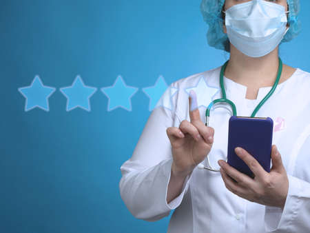 A woman doctor stands on a blue background and holds a smartphone, with the other hand clicks on the virtual rating stars. Assessment of the quality of medicine, ratingの写真素材