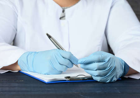 a doctor in a white coat and blue medical gloves sits at the table and writes with a pen, close up