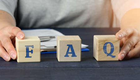 a man sits at a table and holds cubes. inscription FAQ (frequently asked questions) on wooden blocks on a blue background. QA concept, help and tipsの写真素材