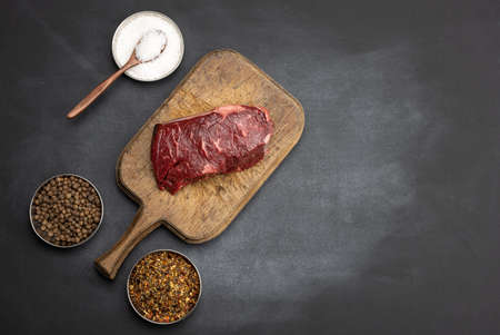raw classic beef steak on a brown wooden board, spice mix, salt on a black table, top viewの写真素材