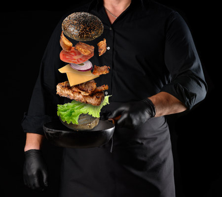 adult man in a black uniform holding a cast iron round frying pan with levitating cheeseburger ingredients: sesame bun, cheese, tomato, onion, meat cutlet, pepperの写真素材