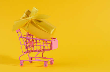 yellow gift box with a silk bow in a miniature metal trolley on a yellow background. Party backdrop, surprise, seasonal saleの写真素材