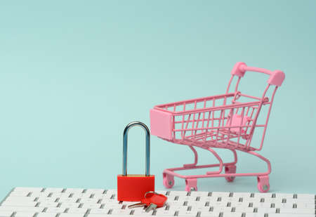 a red metal padlock stands on the keyboard and behind a miniature shopping cart, blue background. Personal data protection concept, secure online shoppingの写真素材
